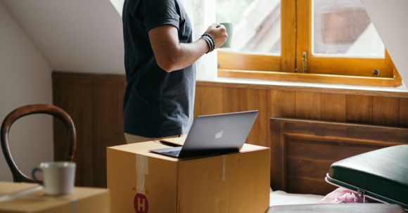 Neighborhood - Side view of young ethnic male owner of newly bought house with cup of coffee in bedroom full of boxes and suitcases standing and looking at neighboring houses through window